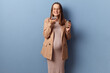 Delighted joyful smiling young adult pregnant woman wearing dress and jacket holding mobile phone recording voice message posing isolated over blue background