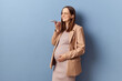 Satisfied delighted young adult pregnant woman wearing dress and jacket posing isolated over blue background recording voice message chatting with her friend online