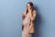 Surprised young adult pregnant woman wearing dress and jacket posing isolated over blue background reading breaking news holding mobile phone browsing internet web pages with shocked content