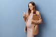 Smiling young adult pregnant businesswoman wearing dress and jacket posing isolated over blue background holding clipboard and using mobile phone for professional chatting