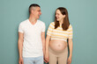 Smiling cheerful pregnant Caucasian young couple standing isolated over light green background wife and husband looking at each other and laughing enjoying happy family moments