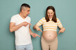 Surprised husband and wife pointing with fingers at big belly looking with excited faces young married pregnant family standing isolated over light green background