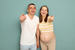 Laughing pregnant Caucasian young couple standing isolated over light green background future parents pointing index fingers at camera choosing or inviting you