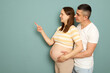 Young married pregnant family standing isolated over light green background man embracing woman pointing at copy space for promotion or advertisement area