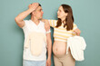 Pregnant Caucasian young couple standing isolated over light green background future mother looking at facepalm exhausted husband tired from buying kid's clothing