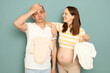 Young married pregnant family choosing clothing for future baby tired husband future father making facepalm gesture standing isolated over light green background