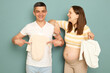 Smiling young married pregnant family standing isolated over light green background future mother and father with tiny bodysuits showing new attired for expecting baby