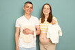 Laughing cheerful joyful pregnant Caucasian young couple standing isolated over light green background holding baby clothing packing bodysuits preparing for childbirth