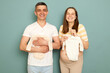 Delighted pregnant Caucasian young couple standing isolated over light green background choosing bodysuits for future baby enjoying pregnancy shopping