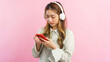 Young women wearing headphone and surfing social media on smartphone isolated on pink background