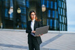 Professional woman working on a laptop outdoors, standing in front of a modern glass building