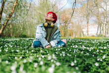 Redhead Woman With Wireless Headphones Sitting Cross-legged On Grass At Park