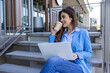 Young woman working on laptop and smiling while sitting at the staircase drinking coffee. Beautiful business woman sitting on the stairs in cafe on stairs and working with laptop on the legs.