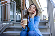 Happy urban woman drinks her takeaway coffee and talk on smartphone. Young woman sits on stairs with tea and holding mobile phone.