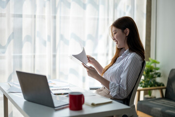 Wall Mural - A woman is sitting at a desk with a laptop and a red mug. She is reading a piece of paper and smiling. Concept of productivity and relaxation