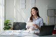 © Wasana - A woman is sitting at a desk with a laptop and a piece of paper. She is smiling and she is happy