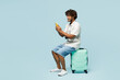 © ViDi Studio - Full body traveler Indian man wears white casual clothes sit on bag use mobile cell phone isolated on plain blue background. Tourist travel abroad in free time rest getaway. Air flight trip concept.