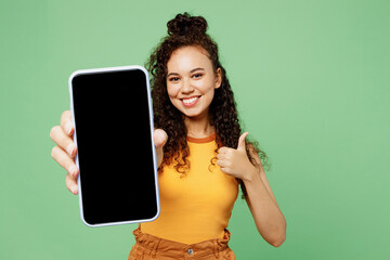 Young smiling woman wear yellow tank shirt top hold in hand use close up mobile cell phone with blank screen workspace area show thumb up isolated on plain pastel green background. Lifestyle concept.
