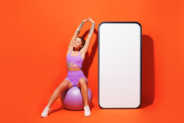 Wall Mural - Full body young fitness trainer woman wear top shorts purple clothes train in home gym big blank screen mobile cell phone sit on fit ball isolated on plain orange background Workout sport fit concept