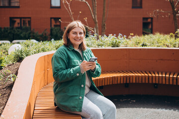 Wall Mural - Close up of woman sitting on the modern bench and using smartphone outdoors. Urban lifestyle concept. Happy 30s woman chatting on cell phone