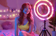 A beautiful redhead teenage girl in pink shorts and yellow top is using ring light for live streaming on her Instagram, in the background there's an aesthetic bedroom with neon lights