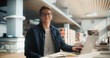 © Gorodenkoff - Confident Young Caucasian Male Student Working On a Laptop in a Modern Library, Surrounded by Books. Smart, Casual Outfit, Glasses, Engaged in Academic Research with a Focused Expression.