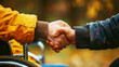 Close-up of a handshake between a person in a wheelchair and a standing person, outdoors on a blurred autumn background.