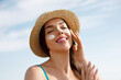 Beautiful young woman at beach applying sunscreen on face and looking at camera. Beauty girl applying suntan lotion at sea. Portrait of happy woman with healthy skin applying sunblock on cheek.