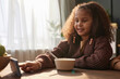 Portrait of teen African American girl using phone at kitchen table during breakfast at home scene lit by sunlight copy space