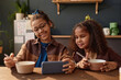 Front view portrait of two smiling African American girls using smartphone at kitchen table in morning scene lit by sunlight