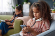 Portrait of young African American girl using smartphone while sitting on couch at home copy space