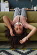 Vertical portrait of little African American girl lying upside down on sofa and looking at camera while having fun at home