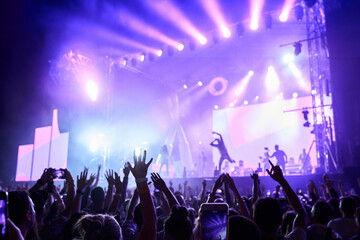 Wall Mural - Silhouettes of excited crowd at music fest, hands raised with lit stage in background. Band performs at night, colorful lights illuminate event. Fans enjoy live concert, moments on smartphones.