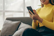 © MrAshi - A young brown-haired woman wearing a yellow shirt sits on a sofa, texting and smiling. Happy model having fun video calling on social media