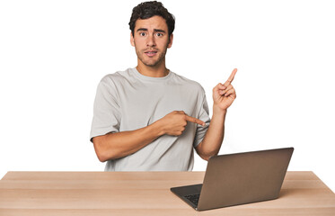 Hispanic young man working on laptop shocked pointing with index fingers to a copy space.