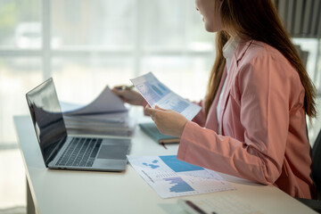 A woman is sitting at a desk with a laptop and a stack of papers. She is looking at the papers and seems to be focused on the information on them. Concept of productivity and concentration