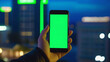 hand holding smartphone with blank green screen against a cityscape. indoor setting and technological elements. Ideal for themes related to app development, smart home technology, or urban living