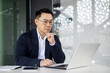 © Liubomir - A professional Asian businessman in a blue suit focuses intently on his laptop screen while sitting at a white table in a sleek, contemporary office space.