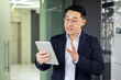 © Liubomir - Confident Asian businessman communicating via a digital tablet in a modern office environment, gesturing with his hand, dressed in a smart business suit.