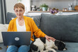 Elderly Caucasian woman sitting on sofa at home with pet French bulldog dog and using laptop
