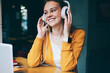 Cheerful hipster girl listening positive playlist via multimedia application and earphones, happy smiling woman enjoying favourite music with high volume in headphones sitting with laptop on front