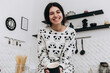 Cheerful woman standing in bright kitchen smiling drinking hot beverage coffee tea, dressed in patterned pajama set. The kitchen counter arranged with ingredients, utensils, atmosphere domestic
