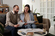Young confident man pointing at financial bill while sitting on couch next to his smiling wife and explaining her data in document