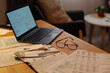 Envelope with dollar banknotes, stack of receipts, pen, open notebook, several financial bills, laptop and eyeglasses on wooden table