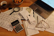 High angle of calculator, envelope with dollar banknotes, magnifying glass, stack of receipts, pen, eyeglasses, financial bills and laptop on table