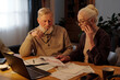 Troubled senior couple sitting by table with financial bills and laptop and looking at unpaid document with past due stamp