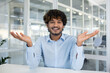 © Liubomir - A cheerful young man with curly hair smiling and gesturing in an office environment, looking camera, giving a relaxed and friendly vibe.