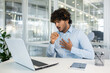 © Liubomir - A young man in a light blue shirt feels a sudden cough while working on his laptop in a modern office setting. Concern and discomfort are evident as he addresses his health at work.
