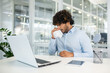 © Liubomir - A young man in a light blue shirt is sitting at his desk in a bright, modern office, visibly sick and using a tissue. This image captures a moment of discomfort in a professional setting.