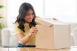 © Prostock-studio - Excited middle eastern woman is standing at a table, opening a box that is placed in front of her. She appears to be focused on the contents of the box, her hands delicately handling the item inside.